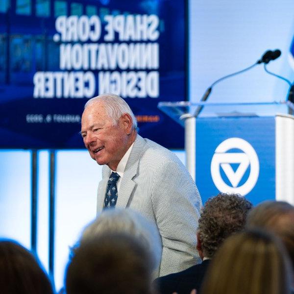Midge Verplank, co-founder of Shape Corp. stands to be recognized at an event. In the background a screen shows Shape Corp. Innovation Design Center