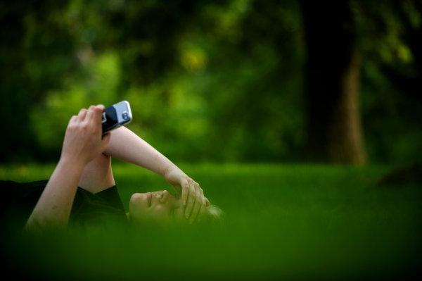 A person lays in the grass with their hand on their head and holding a cell phone